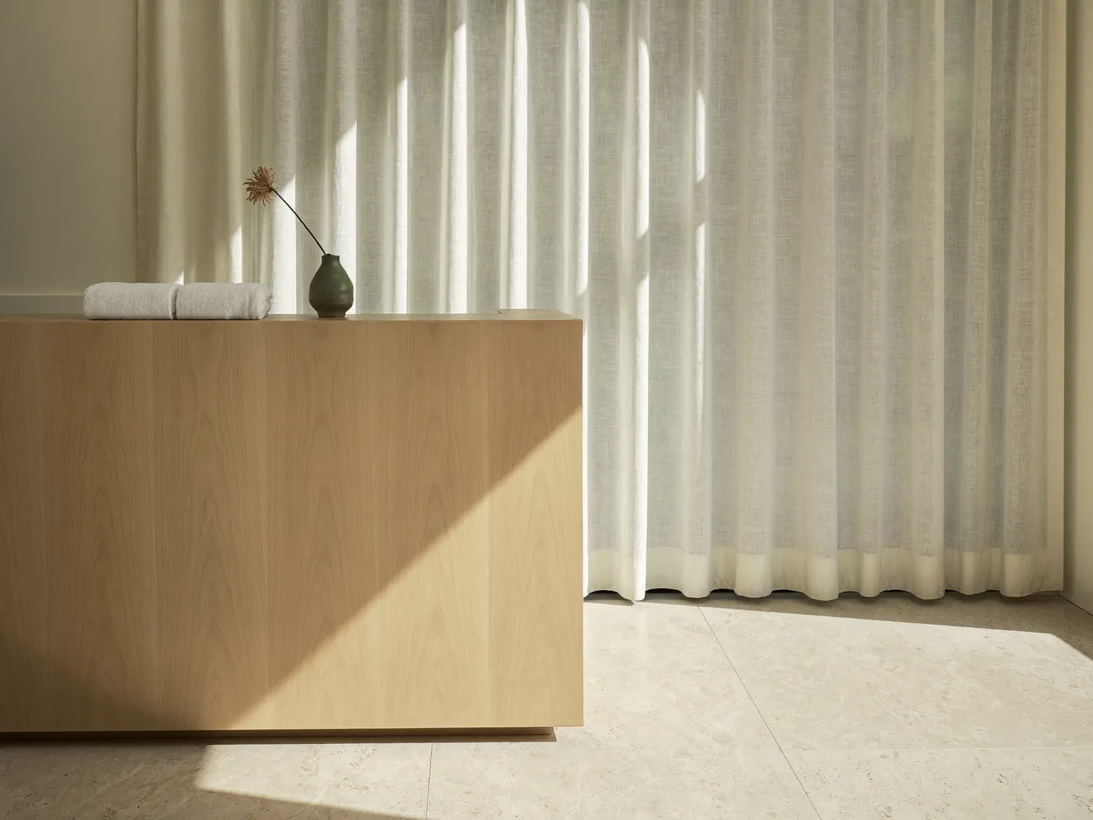 Empfangstresen aus hellem Holz mit gefalteten Tüchern und einer Vase, dahinter ein Vorhang im Morgenlicht